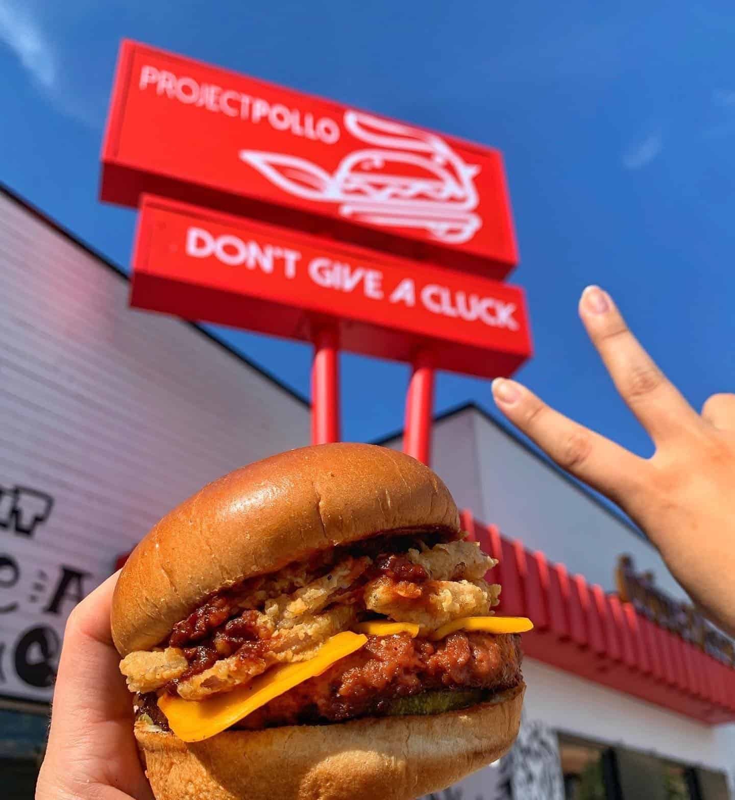 A hand holding a fried chicken sandwich in front of a bright red roadside sign